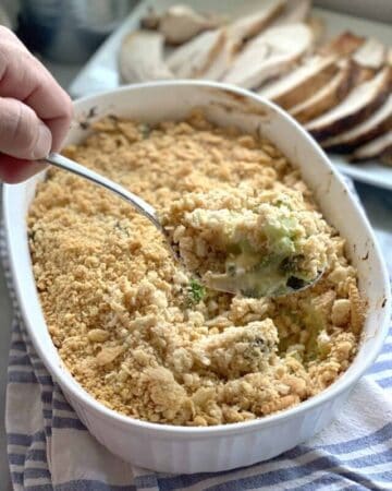 a hand holding a spoon in a casserole dish filled with broccoli casserole topped with ritz crackers