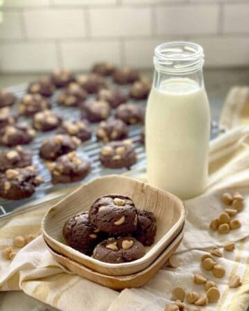 Four chocolate peanut butter chip cookies in a wooden bowl with more cookies on a wire rack and glass of milk in the background