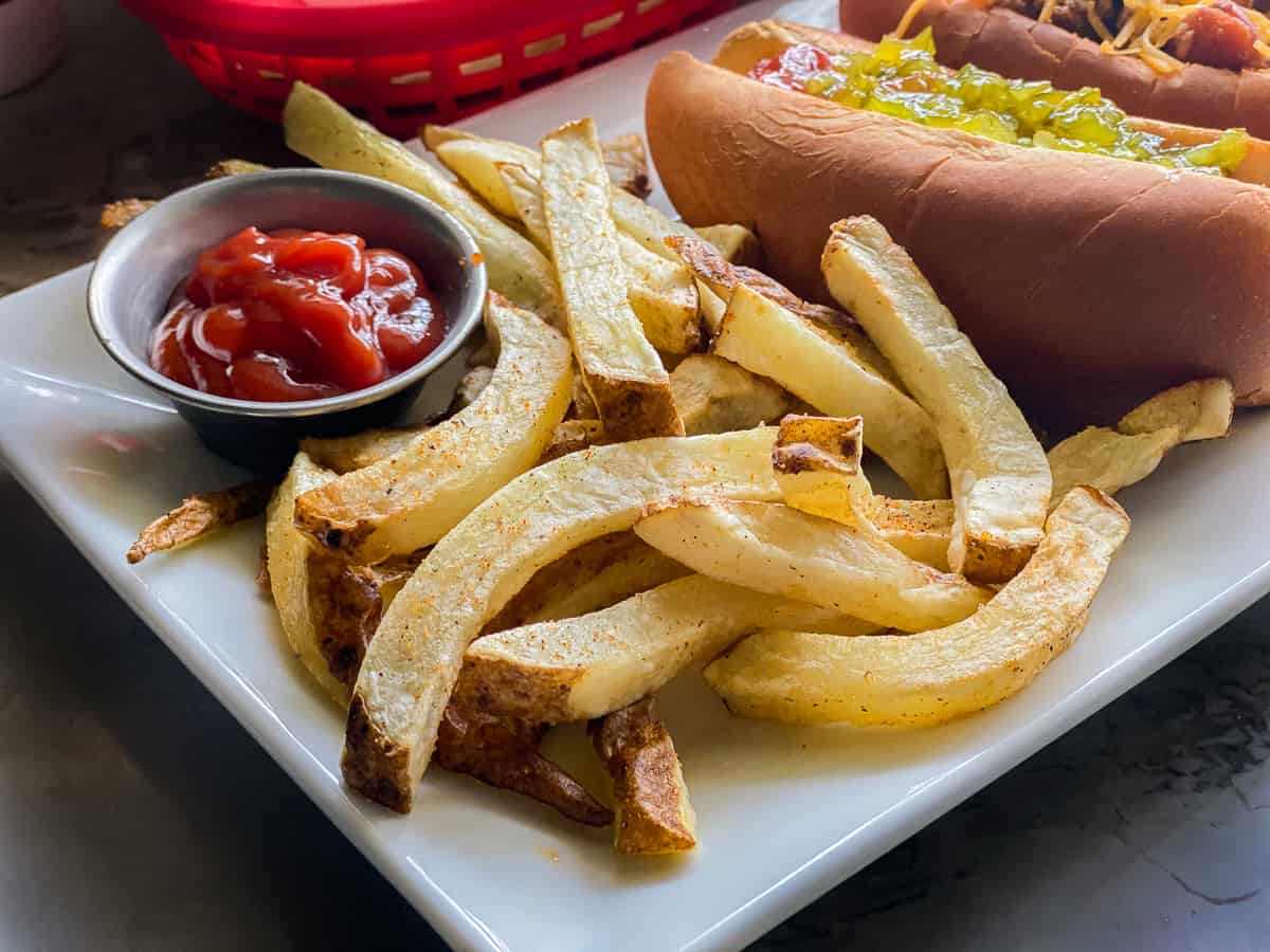 white glass plate with French fries next to gray cup of red ketchup with a hotdog in the background and a red basket