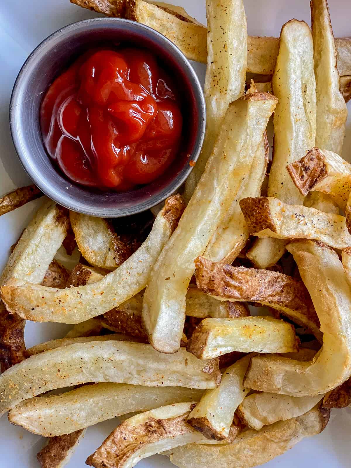 a pile of French fries on a white plate with silver dipping cup with red ketchup