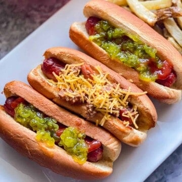 three hotdogs on white plate. far left hotdog with green relish and ketchup middle hotdog with chili topped with yellow shredded cheese, far right hotdog topped with green relish and ketchup. fries in he background