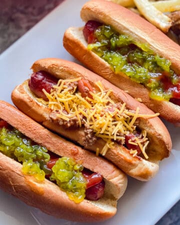 three hotdogs on white plate. far left hotdog with green relish and ketchup middle hotdog with chili topped with yellow shredded cheese, far right hotdog topped with green relish and ketchup. fries in he background