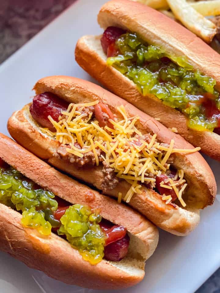 three hotdogs on white plate. far left hotdog with green relish and ketchup middle hotdog with chili topped with yellow shredded cheese, far right hotdog topped with green relish and ketchup. fries in he background