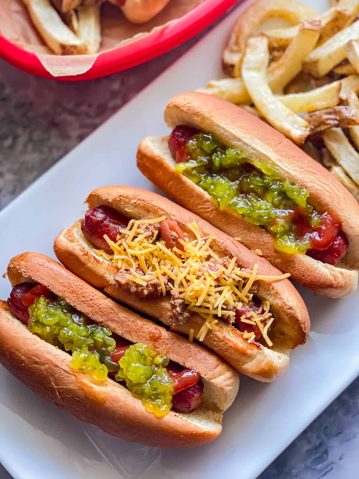 Three hot dogs on a white platter with various toppings and a side of fries