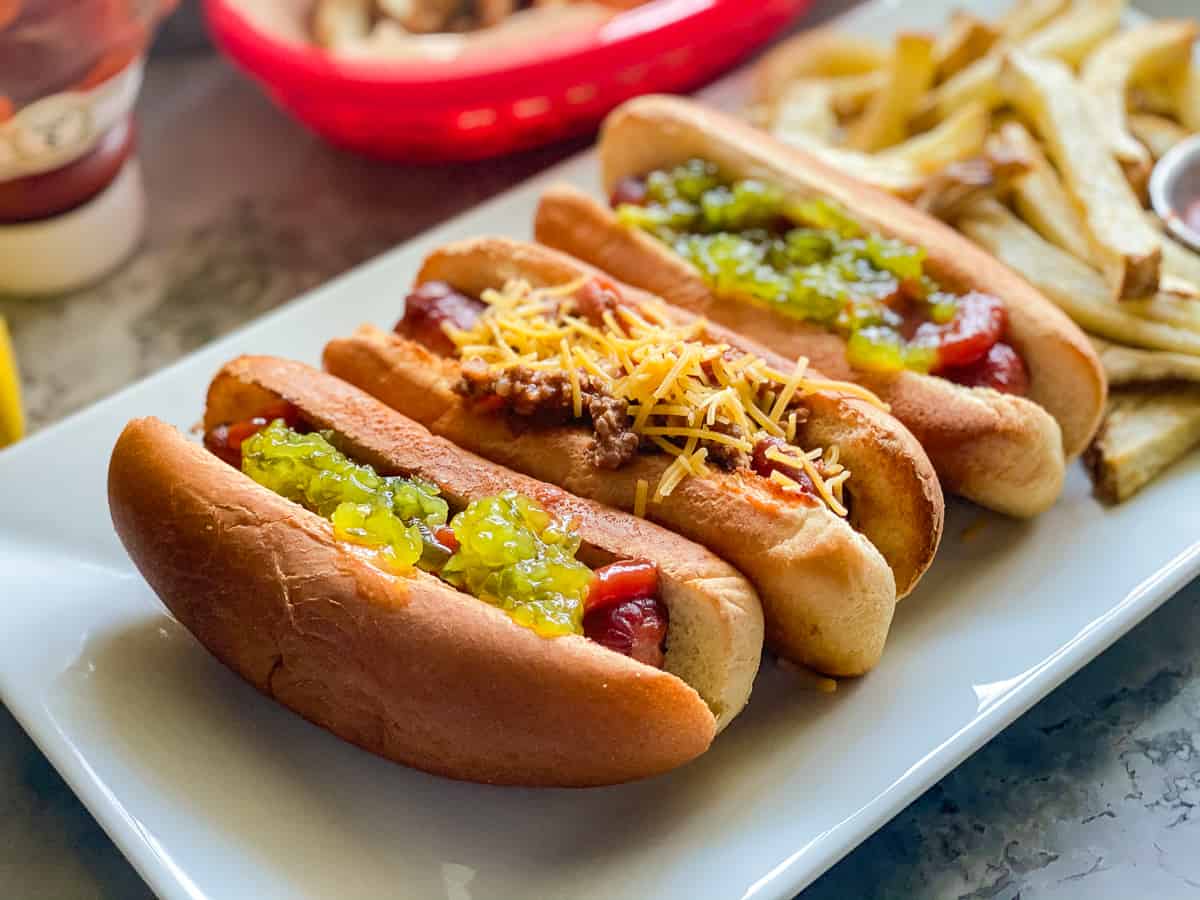 Three hot dogs on a white platter with various toppings and a side of fries
