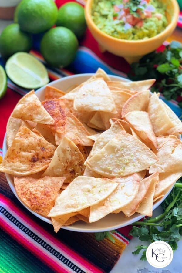 White bowl filled with triangle tortilla chips with guac in background and logo on bottom right corner.
