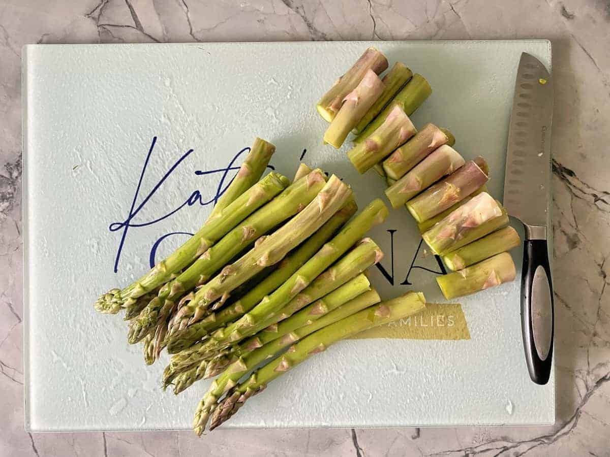 Asparagus with the ends cut next to a silver knife on top of a teal cutting board on a white marble counter