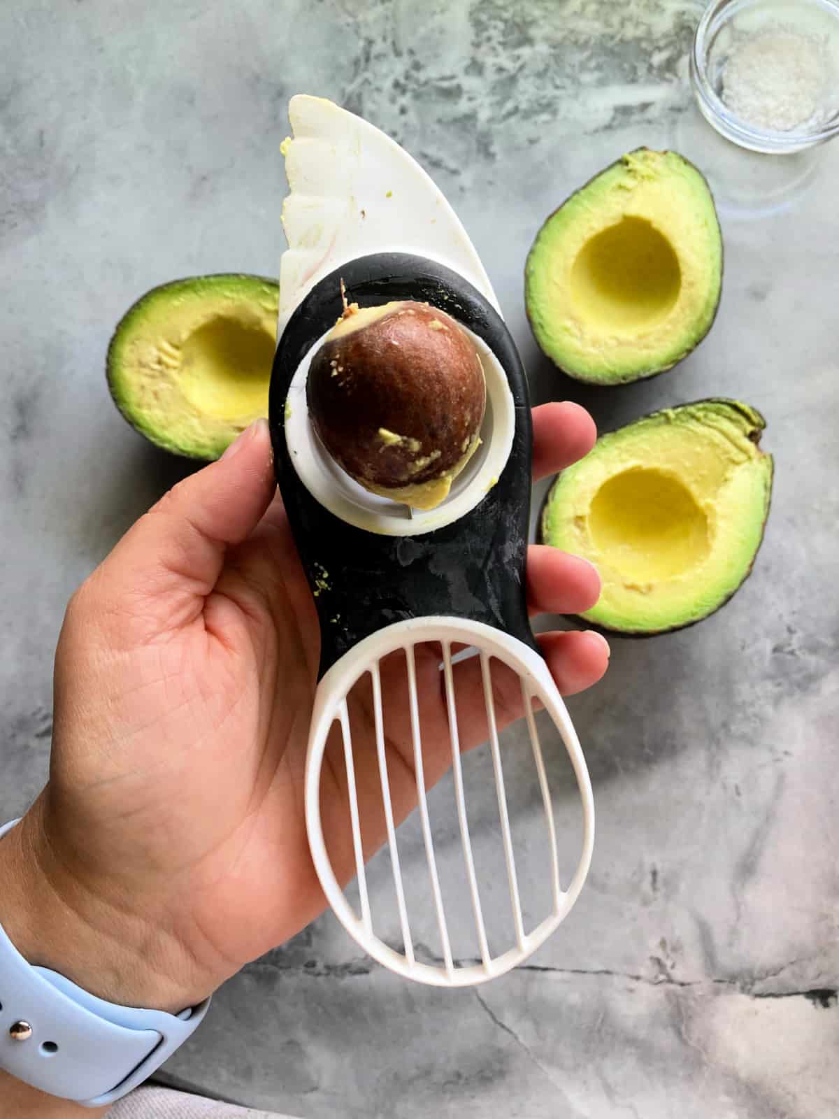 Hand holding avocado slicer with avocado pit in slicer with avocados on counter.