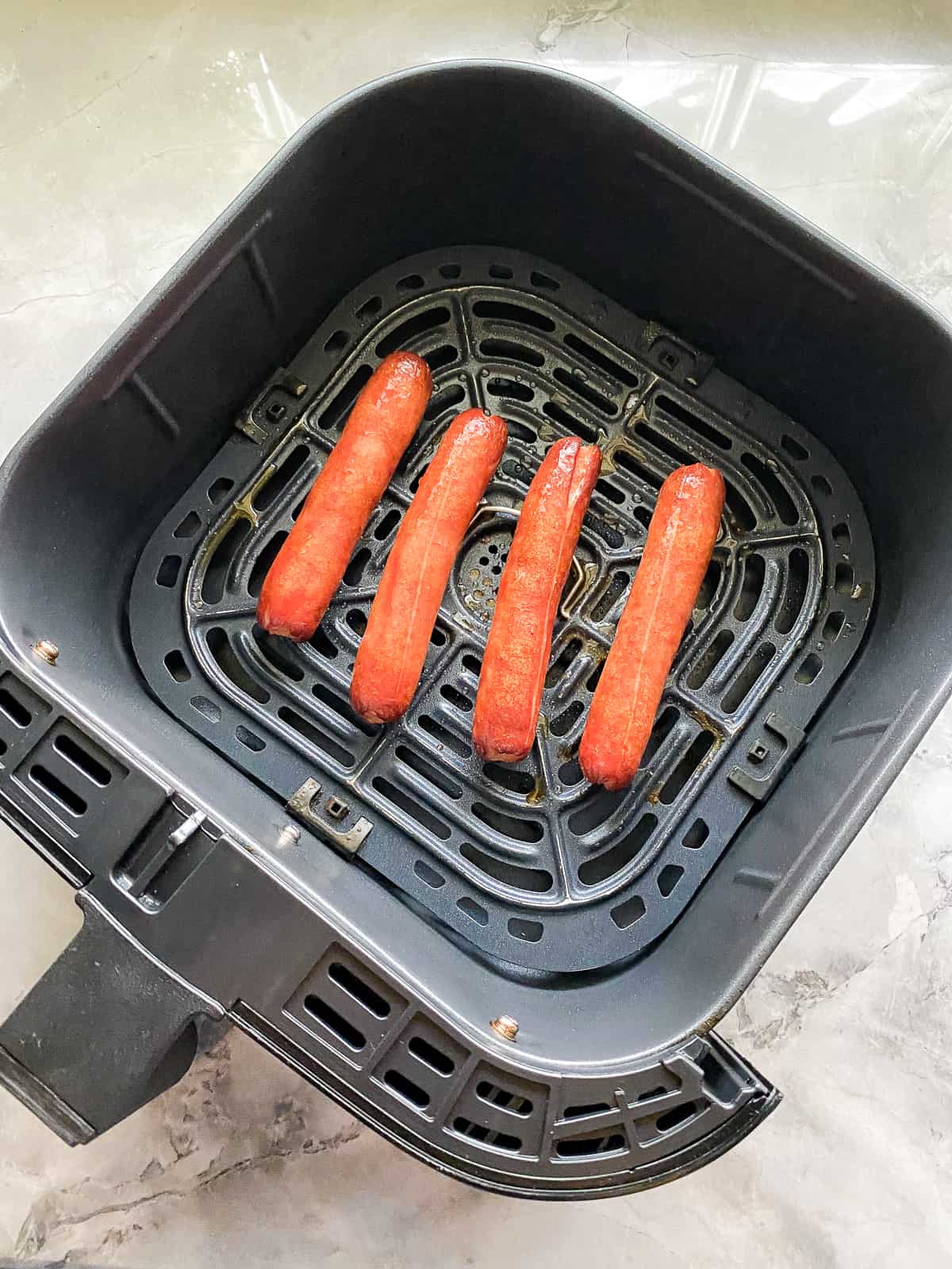 black air fryer on white marble counter with four cooked hotdogs inside