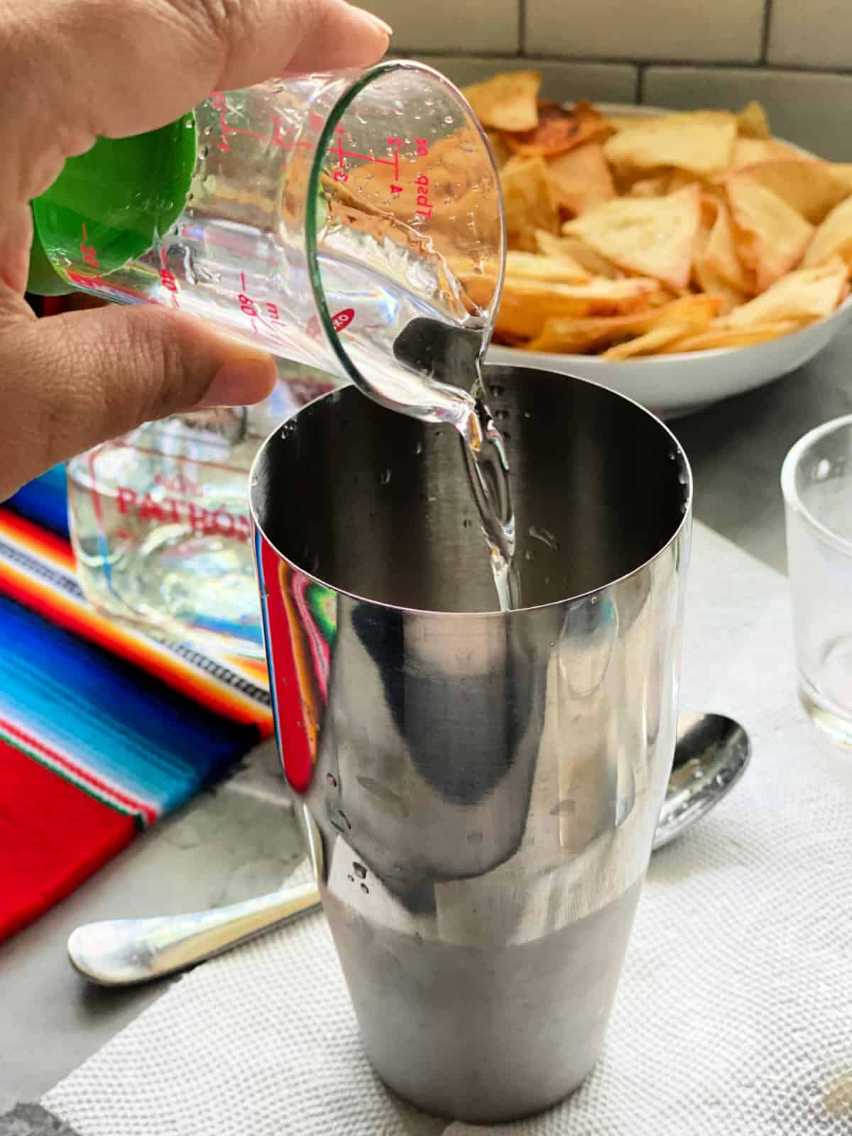 Hand pouring clear liquid into a cocktail shaker.