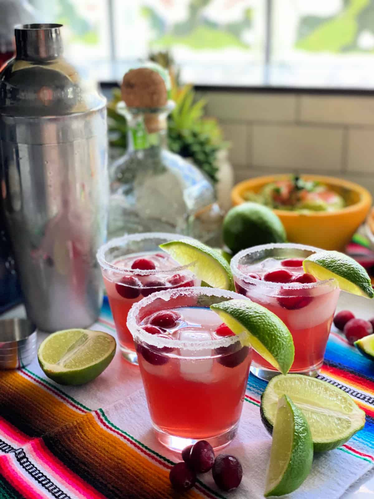 Three glasses with margaritas filled with ice, cranberries, and lime wedges and a shaker in the background.