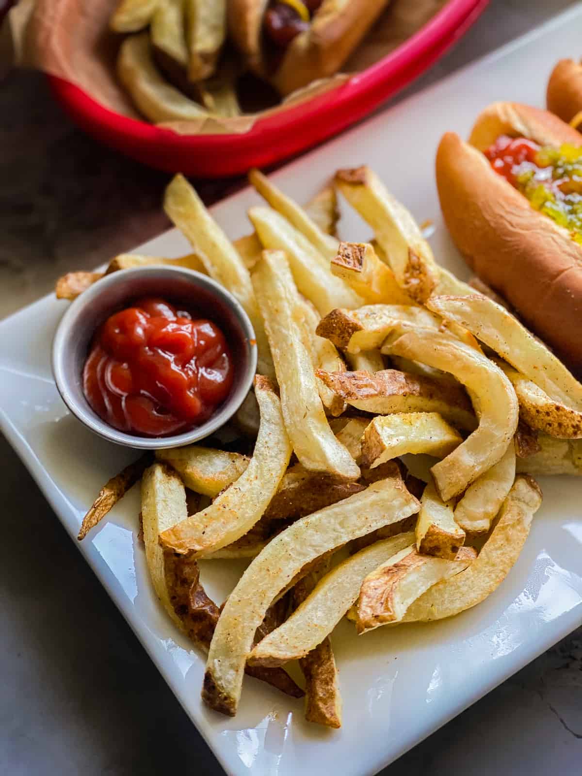 glass white plate with a pile of French fries next to a sliver dipping cup of red ketchup with a hotdog in the background.