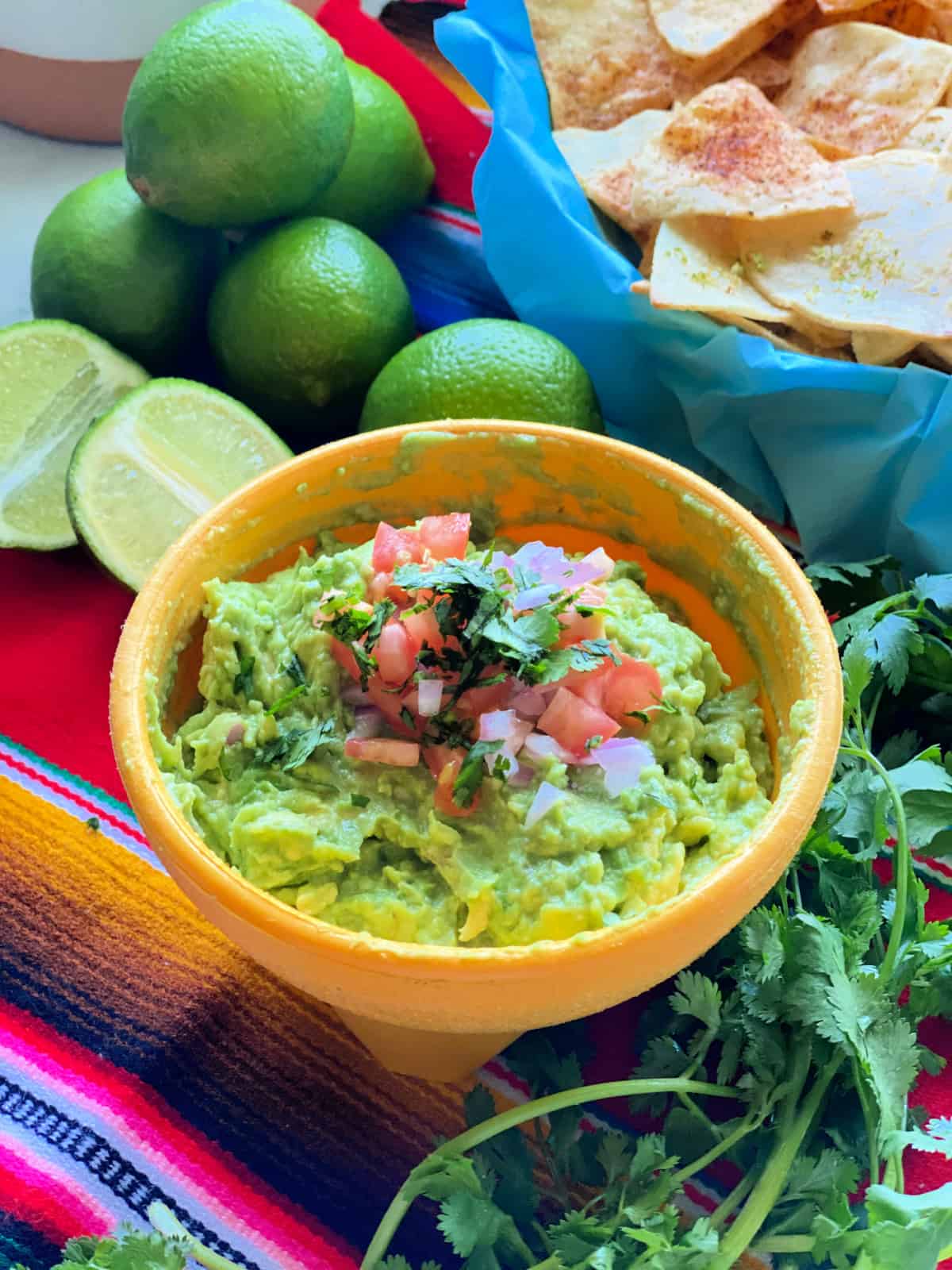 Yellow bowl filled with guacamole tomatoes, red onion, and cilantro, with chips and lime in the background. 