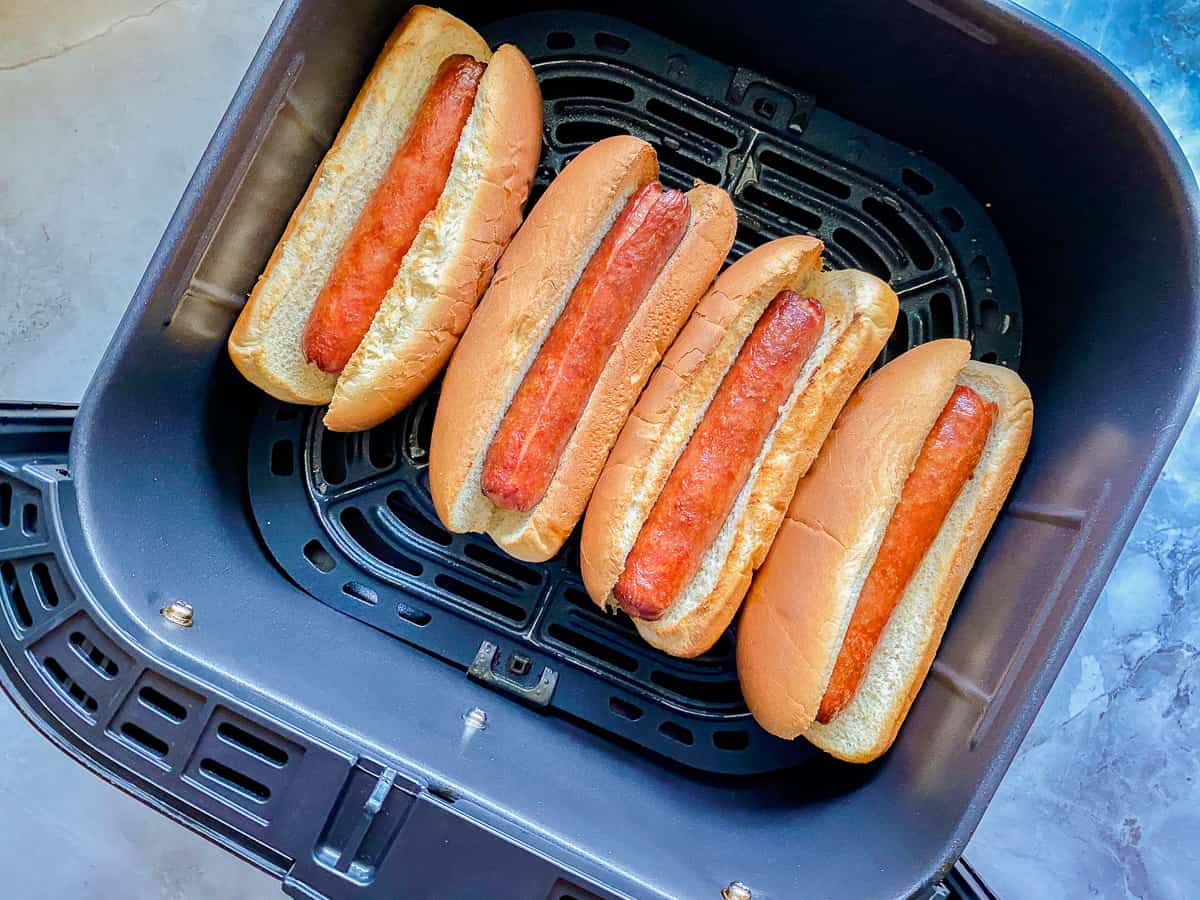 black air Air fryer on white marble counter with four cooked hotdogs in buns inside