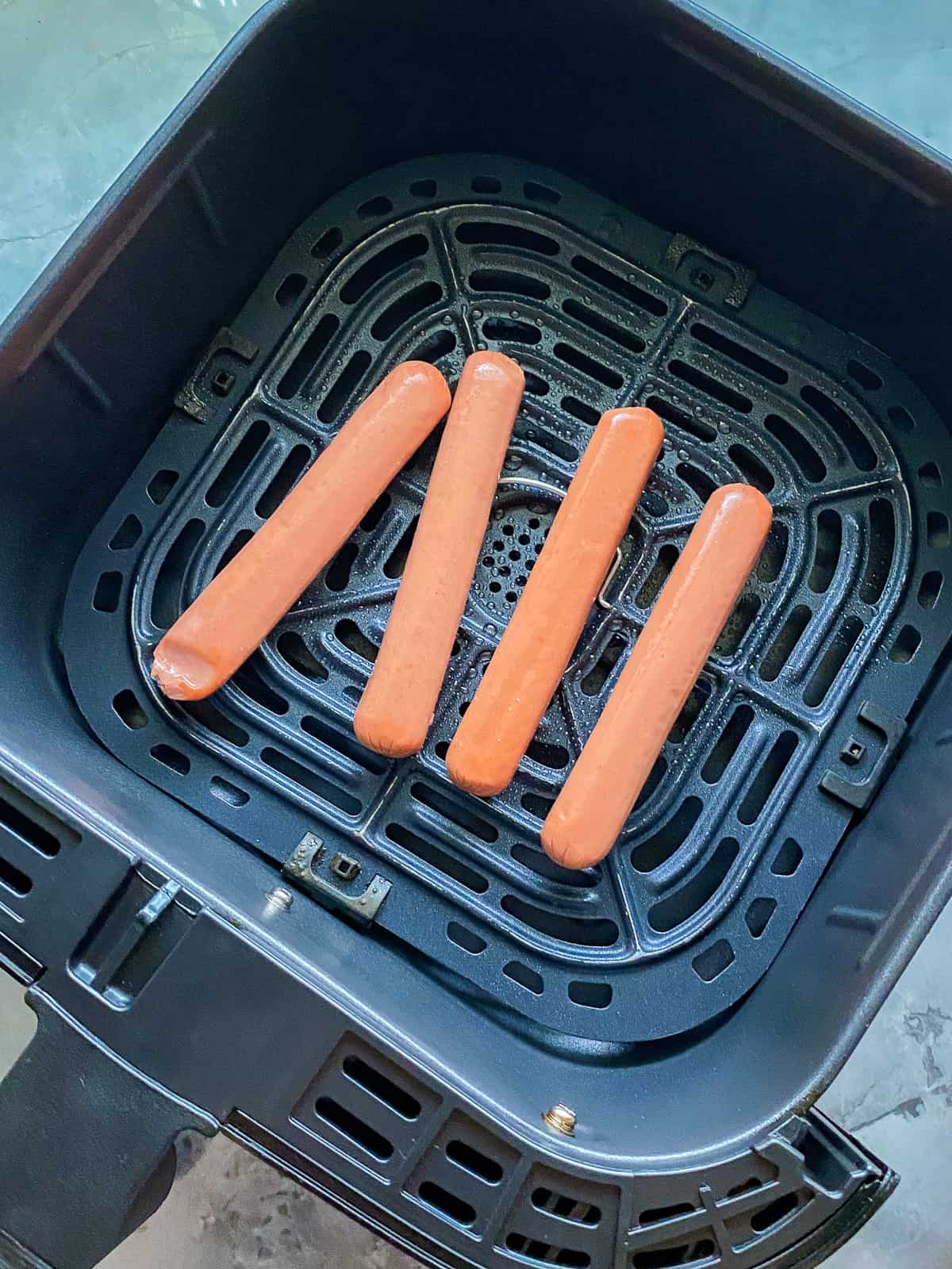 black air fryer on white marble counters with four uncooked hotdogs inside