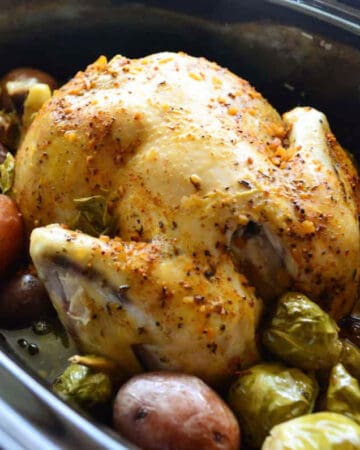 Black slow cooker with a whole chicken resting on top of red potatoes and brussels.