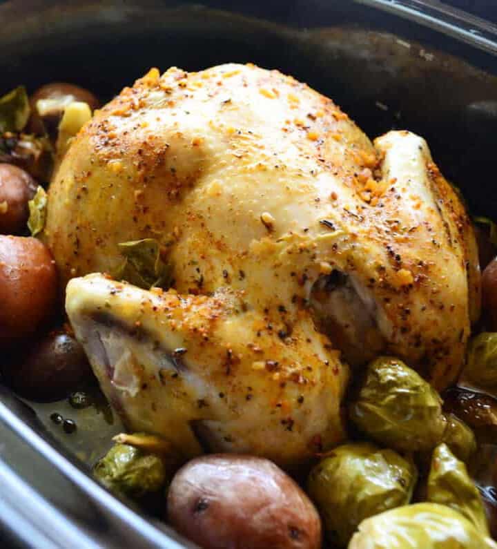 Black slow cooker with a whole chicken resting on top of red potatoes and brussels.