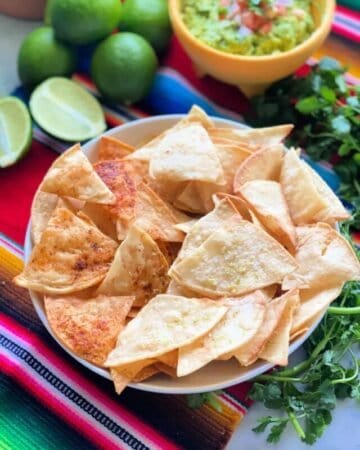 A bowl of tortilla chips surrounded by cilantro, limes, and bowl of guacamole