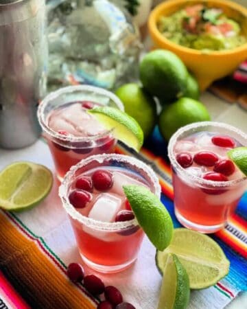 three small glasses of filled with red drink, cranberries and limes