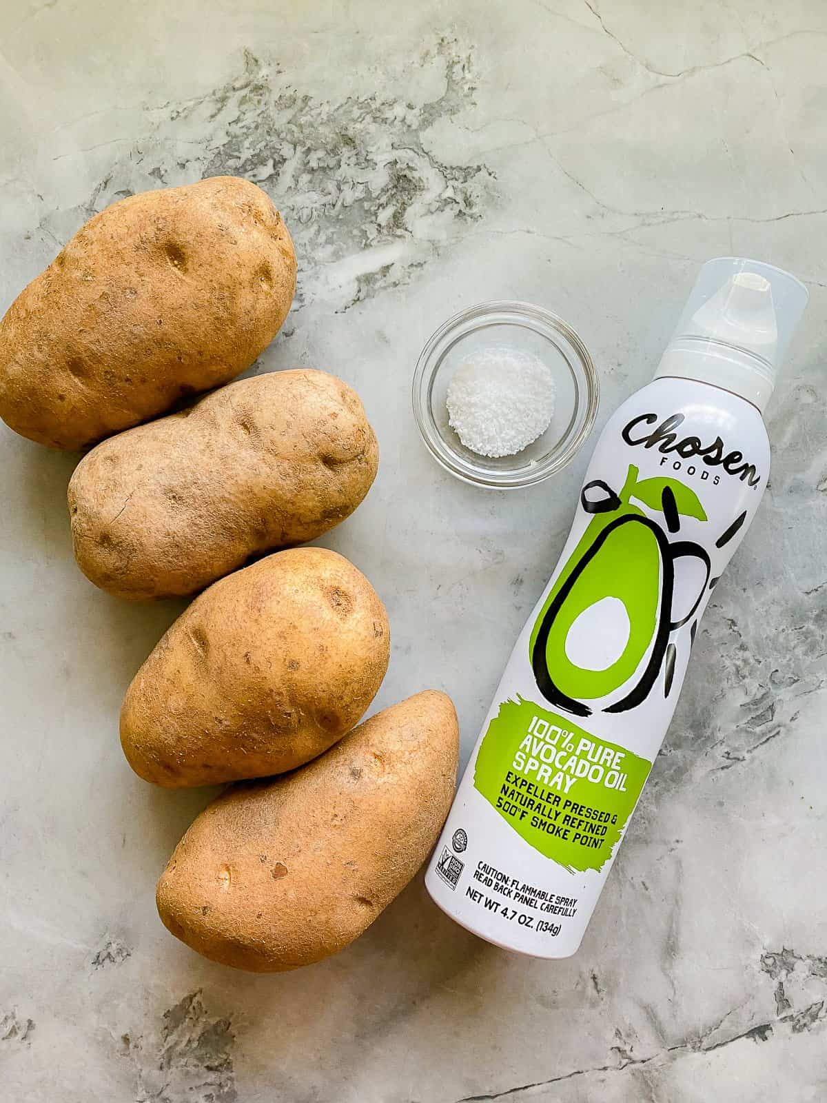 four uncooked potatoes , a small bowl of salt, and a white oil spray can on top of a white marble counter.
