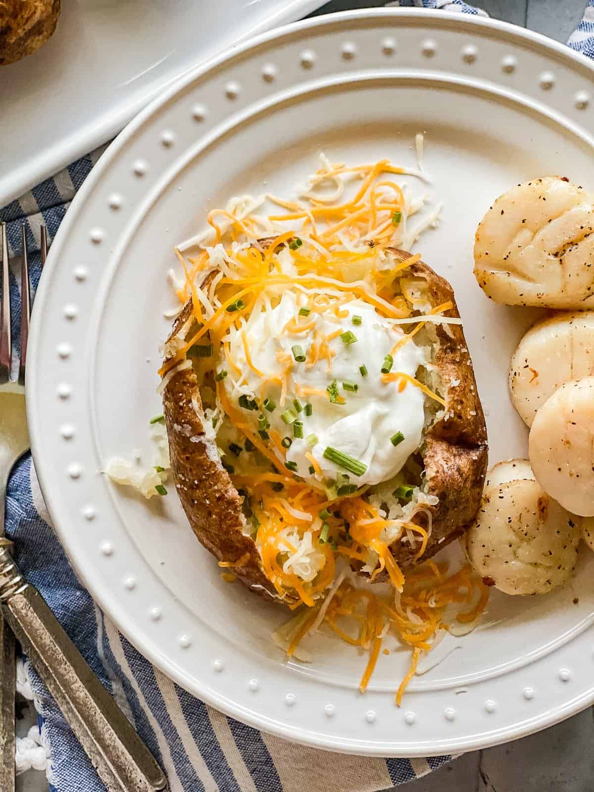 A white plate with a baked potato sliced in the middle and filled with yellow cheese, green scallions, and sour cream. Next to the potato are five scallops.