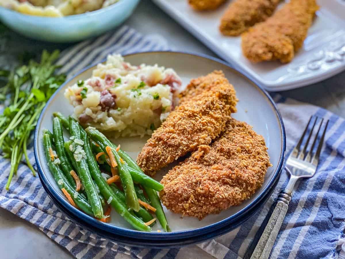 white plate with blue rim with green beans with yellow cheese next to a golden brown chicken tender next to mashed potatoes with red bacon pieces.
