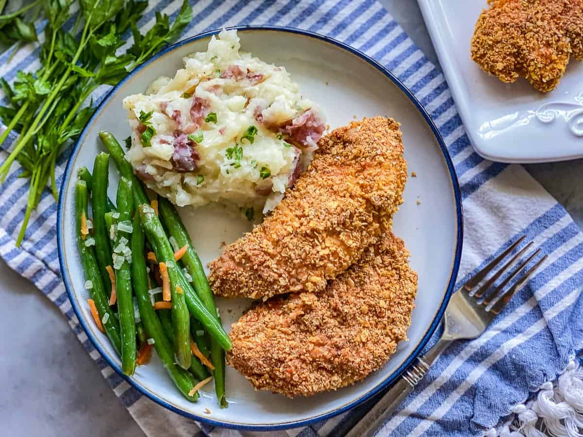 white plate with a silver fork next to plate. with blue rim with green beans with yellow cheese next to a golden brown chicken tender next to mashed potatoes with red bacon pieces.