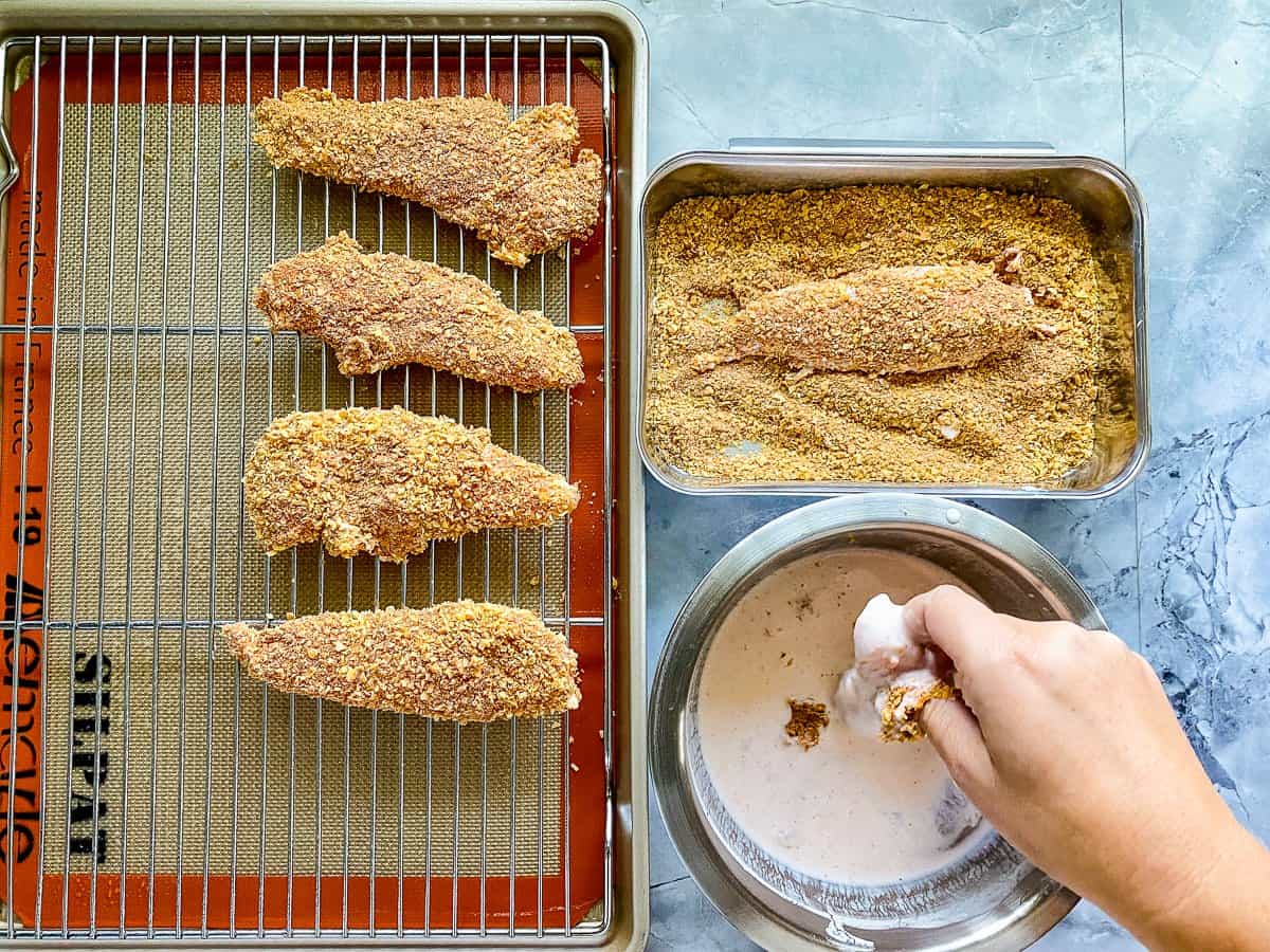 silver pan with red trim and silver rack on top with four pieces of chicken tenders covered in golden frosted flakes next to a silver bowl of pink sauce with raw chicken inside.