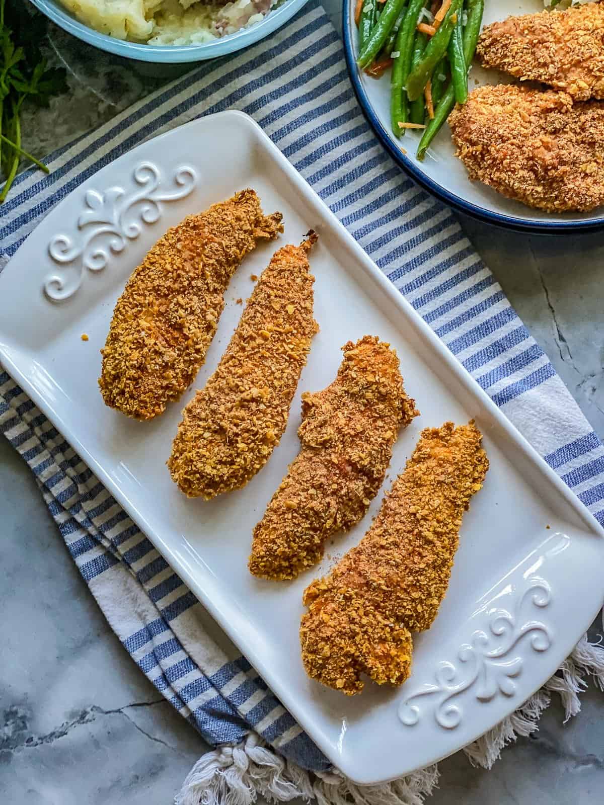 Crunchy-Baked Chicken Strips Rectangular white plate with four chicken tenders, with a second plate on top with green beans.