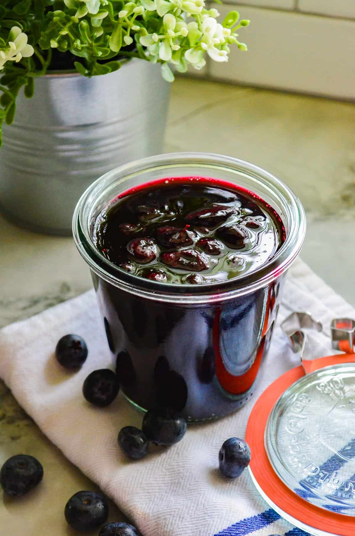 clear cup filled Blueberry Compote on a white napkin with blueberries on it, with a red and clear lid, with a silver pot with a green plant on the top left side.