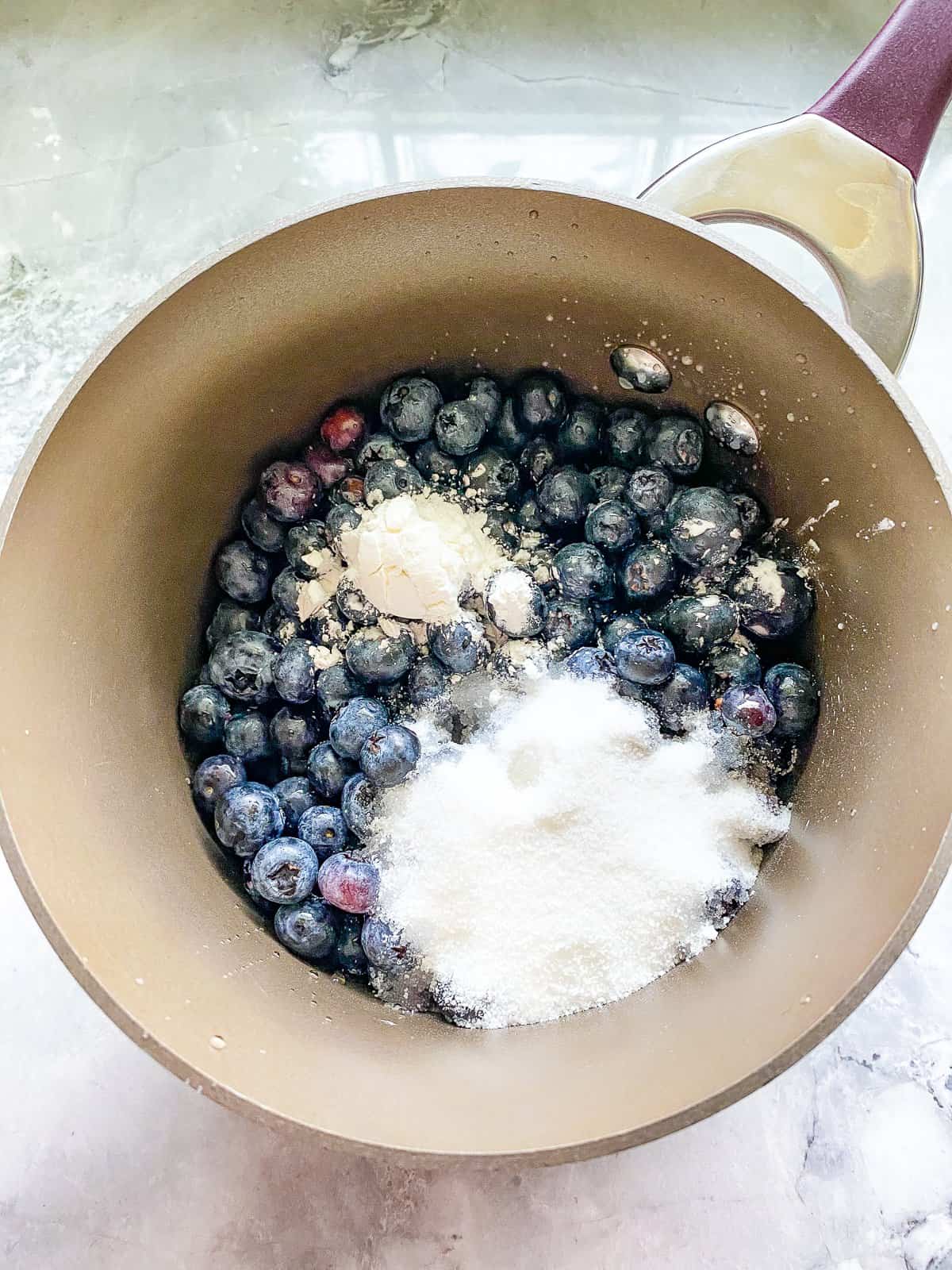 white counter with a pot with a pink handle filled with blue blueberries with white sugar on top.