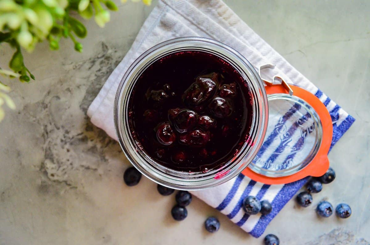 Top view of clear cup filled Blueberry Compote on a white napkin with thirteen blueberries on it, with a red and clear lid, and a green plant on the top left corner.