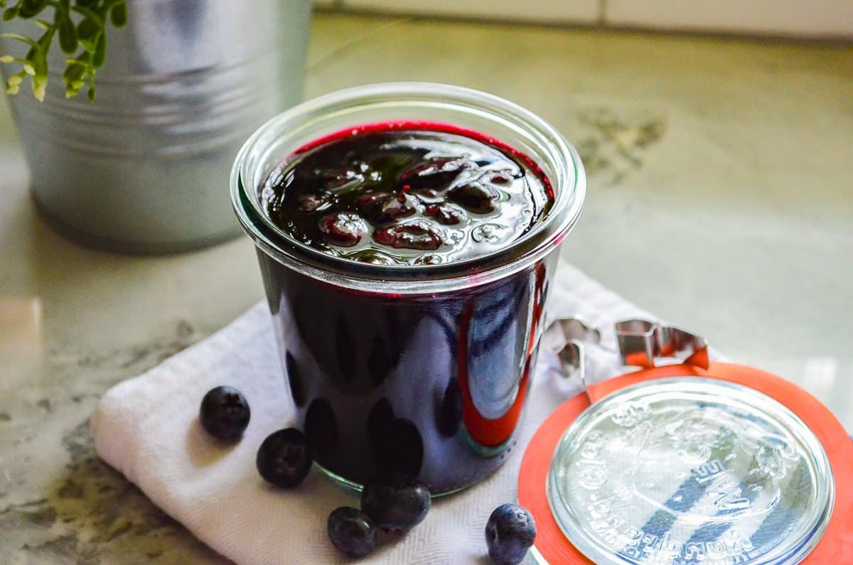 clear cup filled Blueberry Compote on a white napkin with five blueberries on it, with a red and clear lid,