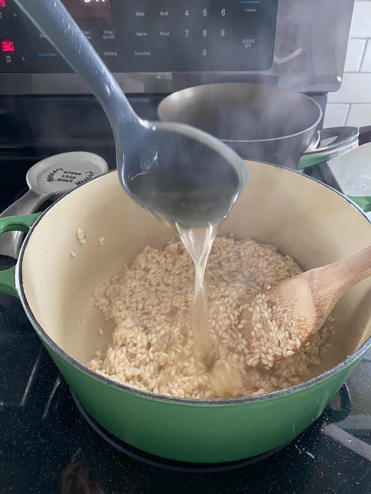 Black ladle pouring hot liquid into a large green stock pot with arborio rice in it. 