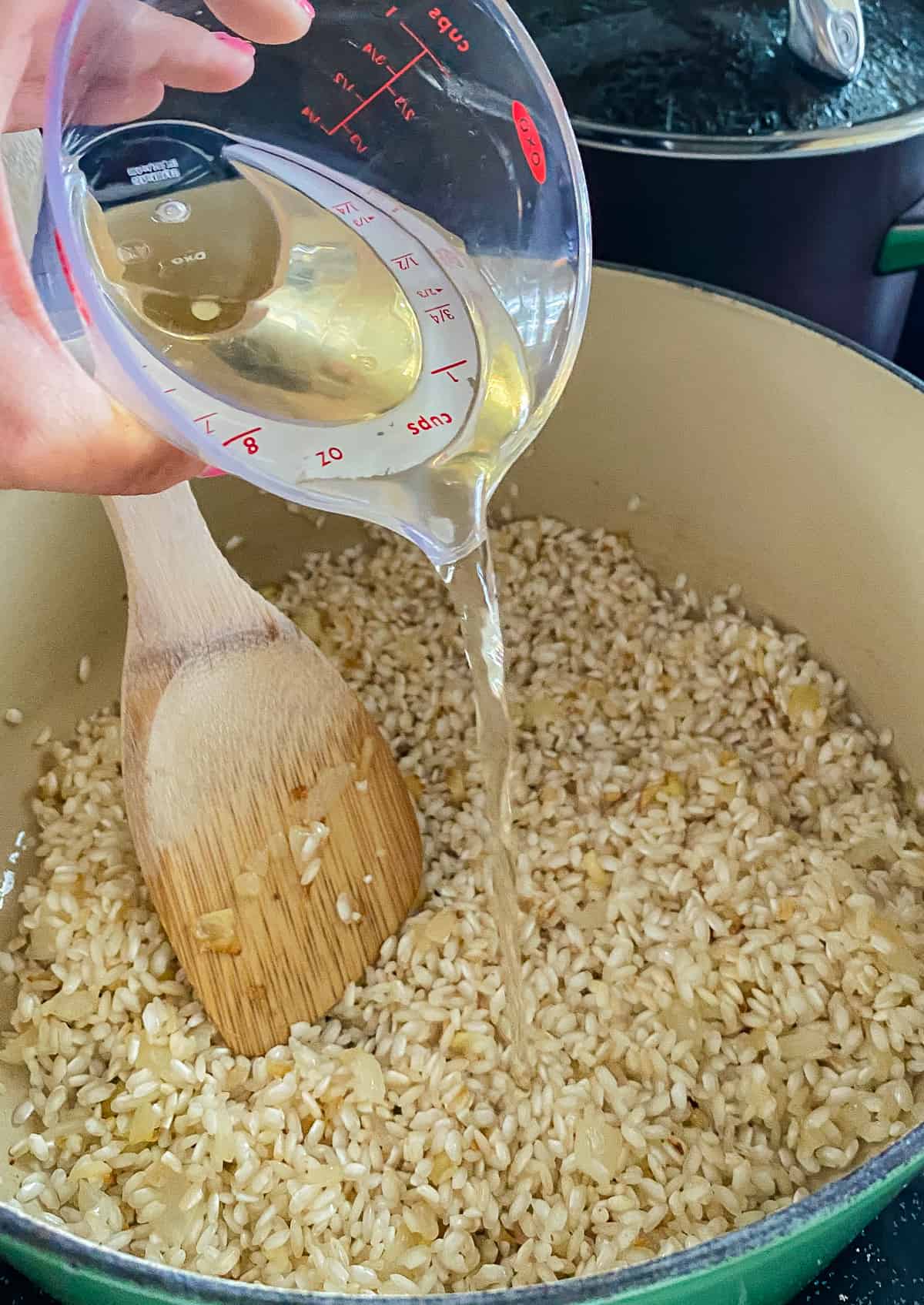 Handing pouring clear liquid into a pot of raw arborio rice with a wood spoon in the pot. 