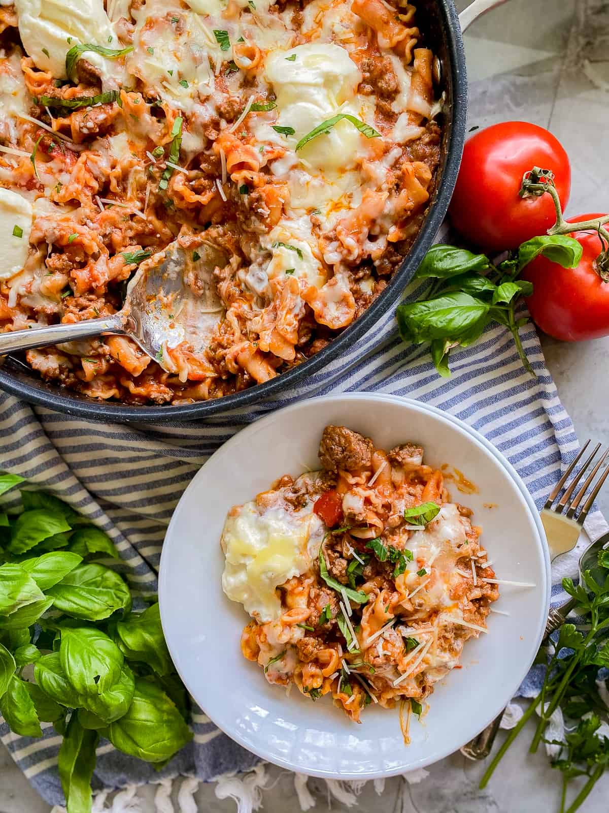 Lasagna in a skillet with a white bowl of lasagna on a striped cloth napkin, basil leaves, and tomatoes on the vine. 