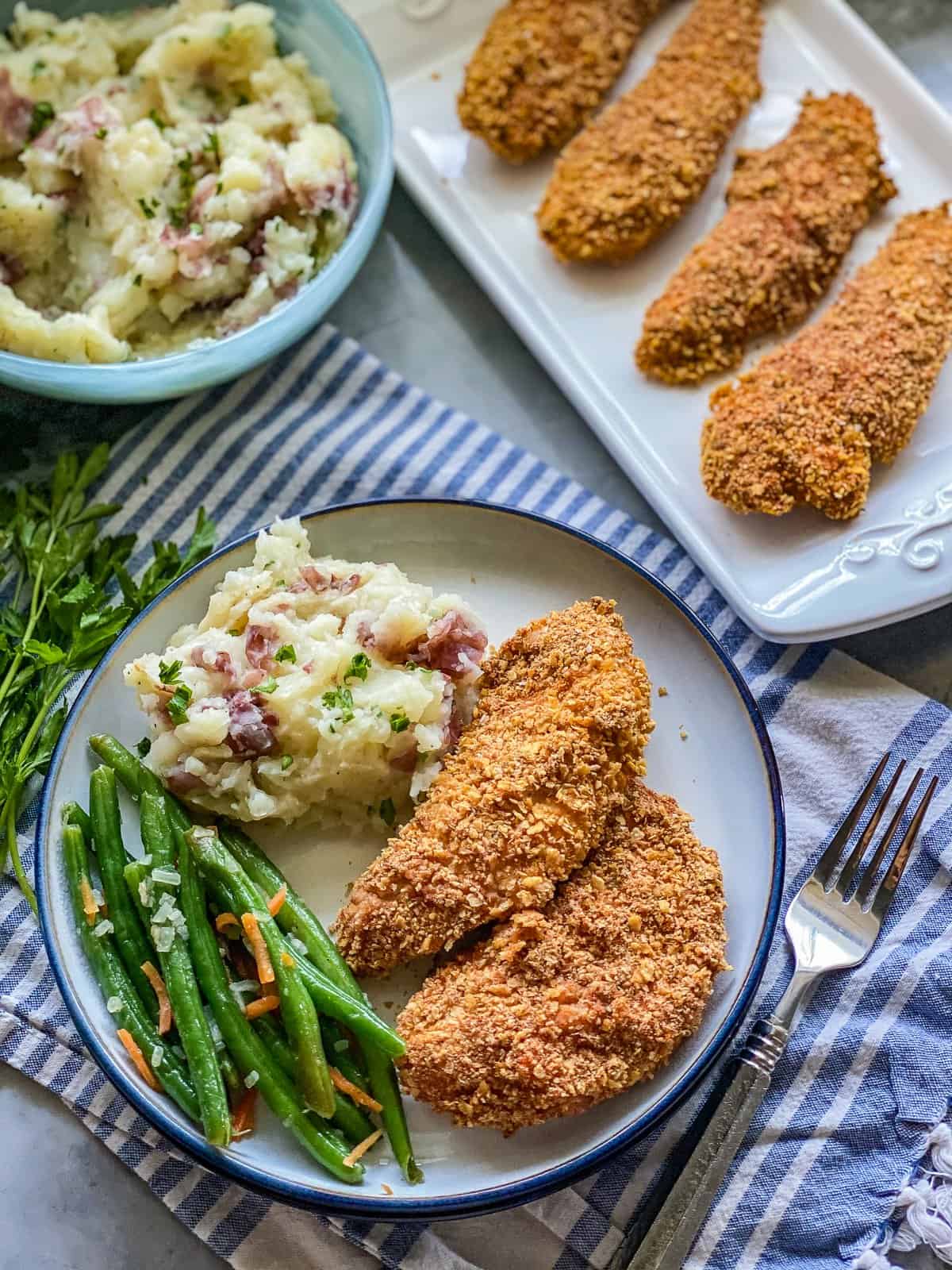 Crunchy-Baked Chicken Strips a white plate with two pieces of chicken tenders next to green beans with yellow cheese flakes and mashed potatoes with green scallion and red bacon bits.