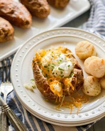 A white plate with a baked potato sliced in the middle and filled with yellow cheese, green scallions, and sour cream. Next to the potato are five scallops.