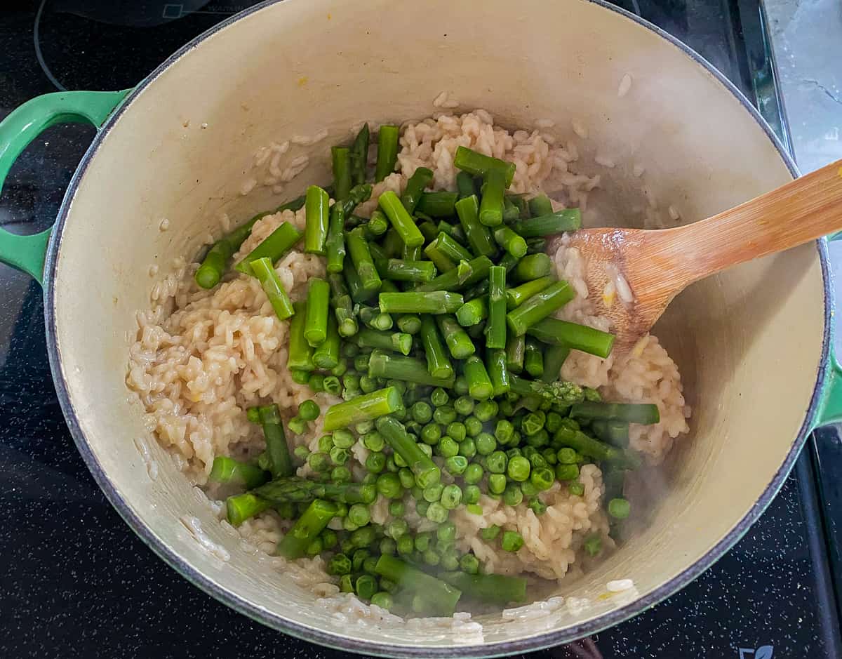Green pot with peas and asparagus on top of arborio rice with a wooden spoon.