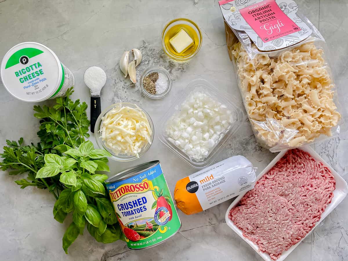 ingredients on counter; basil, parsley, crushed tomatoes, onions, sausage, ground beef, pasta, seasonings, oil, and ricotta cheese.