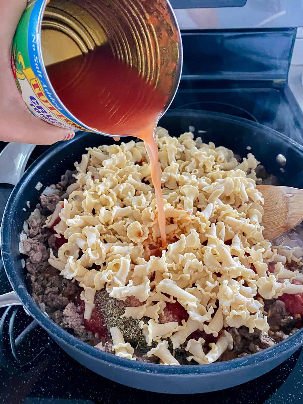 a can with red sauce being poured in the black skillet with brown ground beef and yellow lasagna pasta.