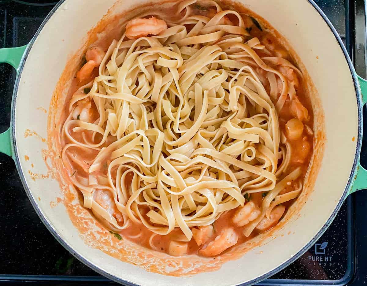 white pot with teal handles on stove filled with yellow pasta noodles, red sauce and shrimp and scallops.