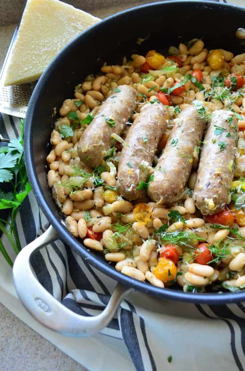 5 sausages in skillet over beans with basil and cheese next to parsley on tablecloth.