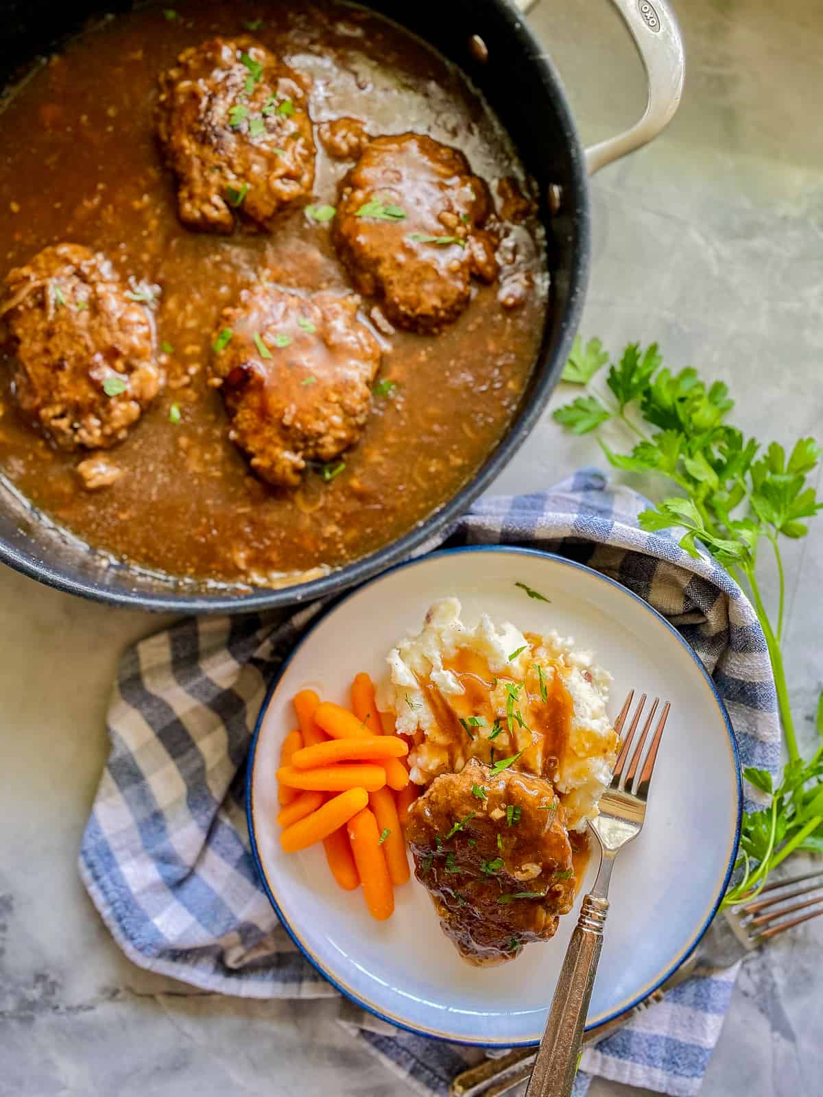 white plate with blue trim with white mash potatoes, orange carrots, brown steak with gravy with silver fork. Next to a big black skillet with cooked steaks with brown gravy inside.