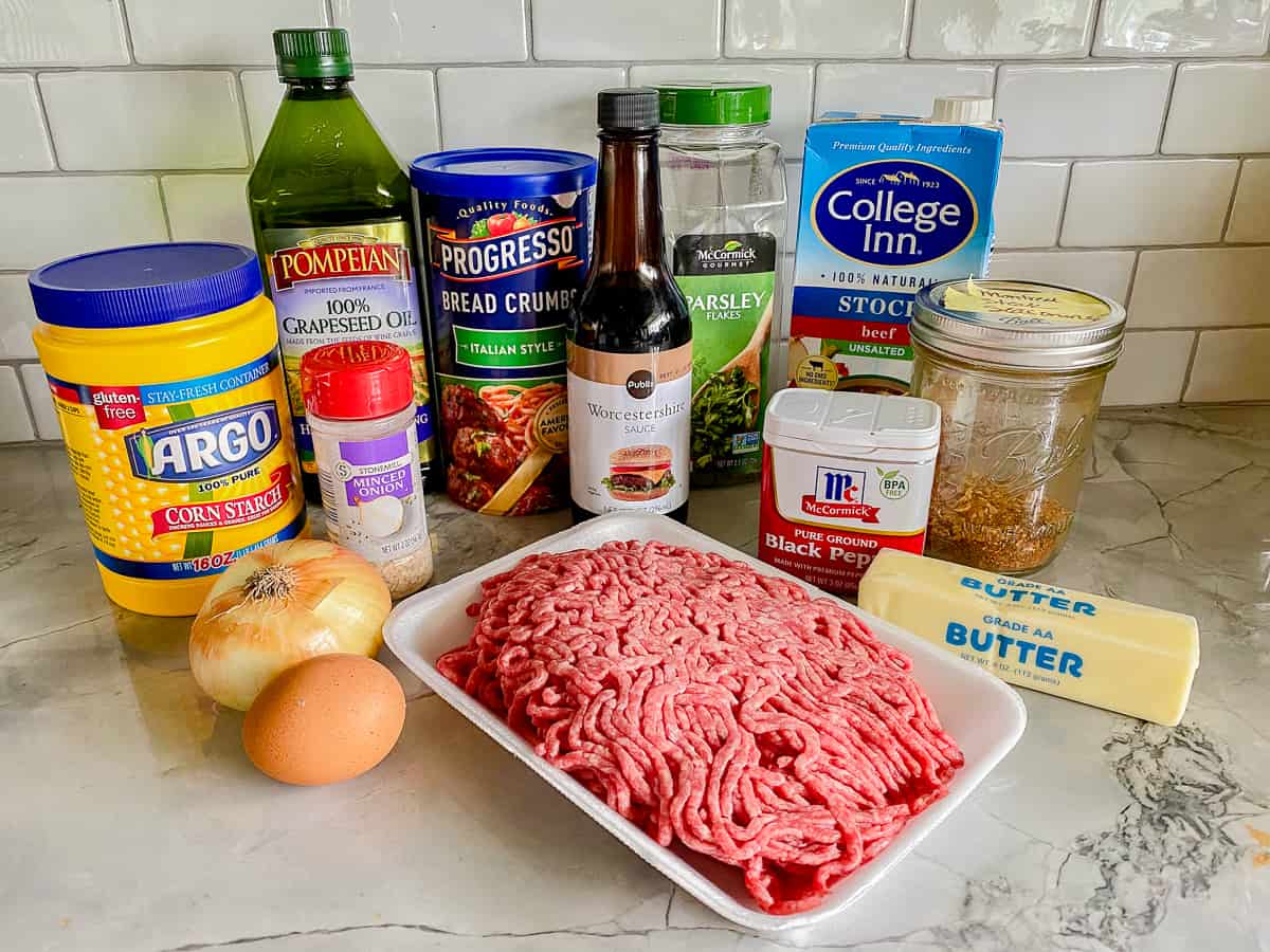 uncooked beef, butter, brown egg, yellow onion, black pepper container, ingredients on a white marble counter.