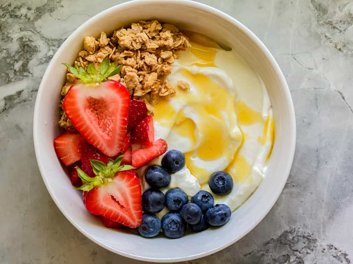 White bowl filled with yogurt, honey, bleuberries, strawberries, and granola on a marble countertop.