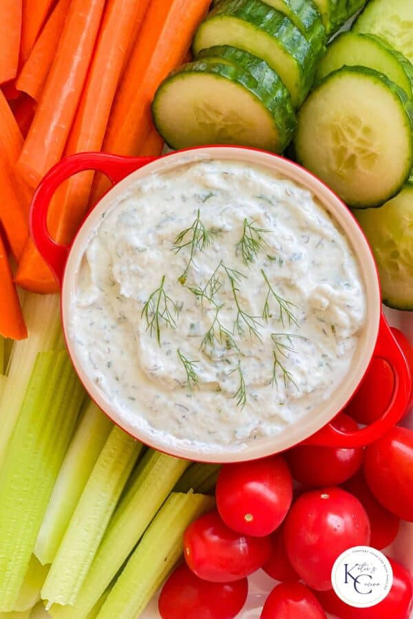 White dip in a red crock with veggies around it and a logo on the bottom right corner.