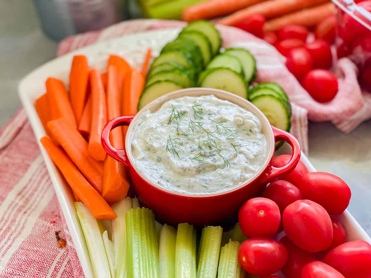 Red crock with white dip with dill on top with carrots, celery, cucumbers, and tomatoes.