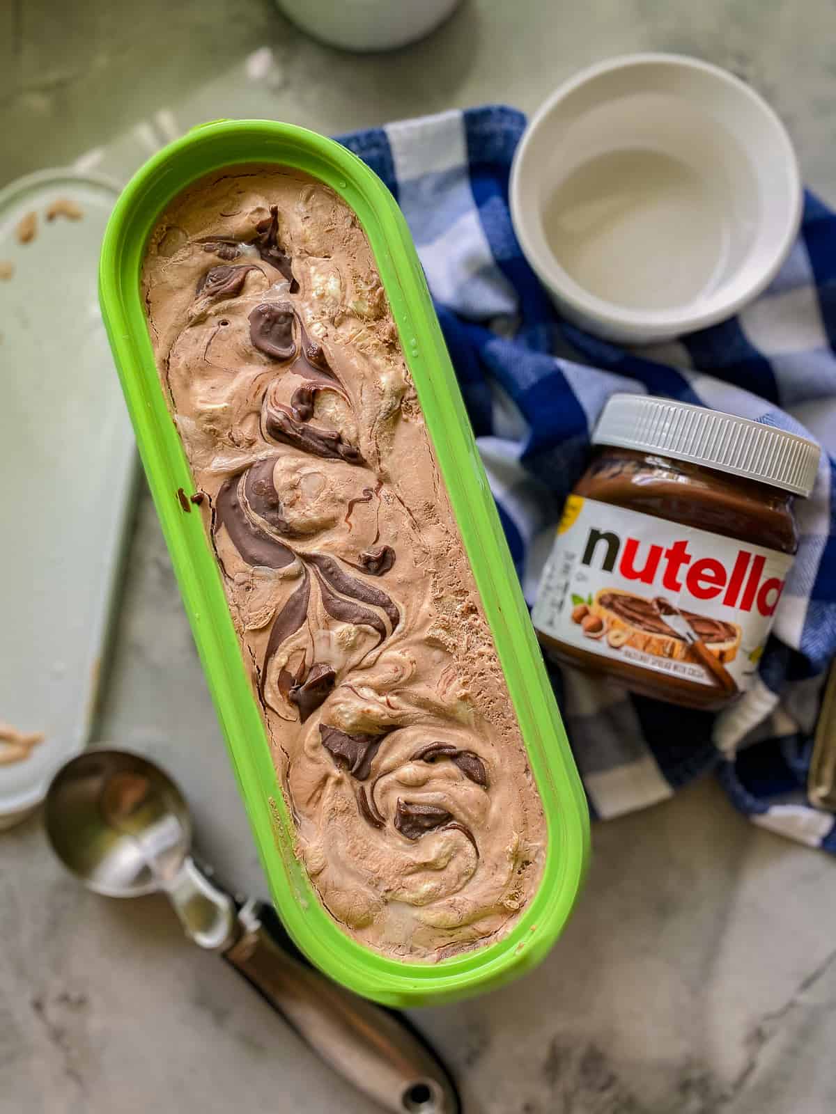 Green oval container with chocolate ice cream, ice cream scoop, and nutella container.