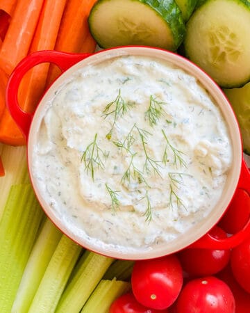 Red crock with white dip surrounded by veggies.